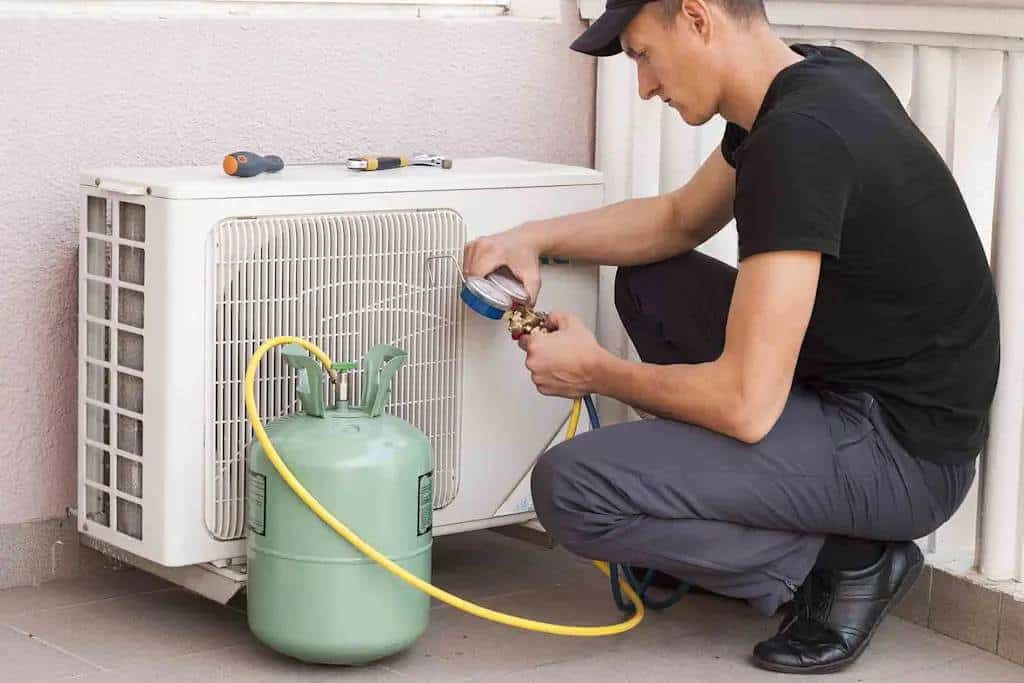 Technician kneeling beside an outdoor air conditioning condenser unit, connecting a refrigerant cylinder with hoses for regassing