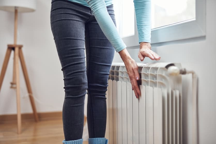 Woman adjusting a white wall mounted radiator heater beneath a window in a living room.